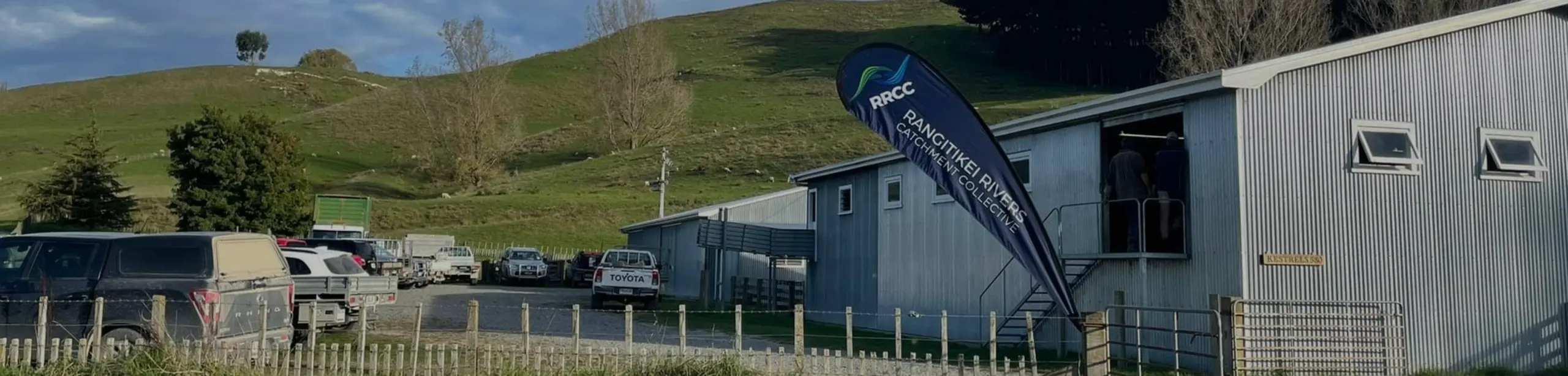A rural New Zealand farm setting with rolling green hills and trees in the background, several utes and farm vehicles parked near a corrugated metal building, and a large flag outside reading RRCC Rangitikei Rivers Catchment Collective.